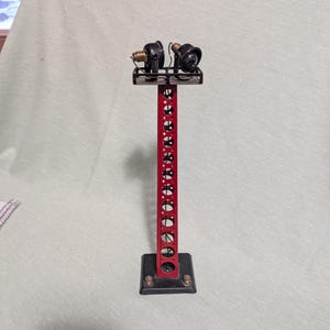May include: A vintage model train signal with a red tower and black base. The tower has a series of circular cutouts and supports two black lamps. The base is square with four bolts.