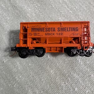May include: An orange model train car with the text "MINNESOTA SMELTING" printed on the side. The model has black wheels and is set against a gray background. The text "MSCX 122" is also visible.