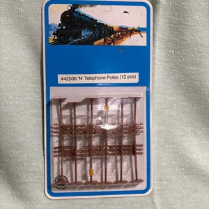 May include: Package of twelve brown plastic model telephone poles for a miniature train set. The packaging is blue with an illustration of a train and the text "#42506 'N' Telephone Poles (12 pcs)".