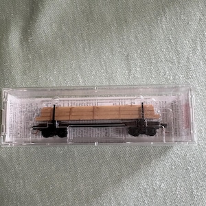 May include: A miniature model train flatcar carrying a load of wooden planks. The flatcar is black with four wheels and is encased in a clear plastic box. The wooden planks are light brown and stacked neatly on the flatcar.