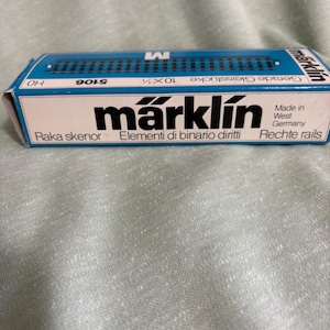 May include: A blue and white Märklin box containing model train rails. The box has the text "Märklin" in a bold font, along with the text "Made in West Germany" and "5106". The box is rectangular and has a blue top.