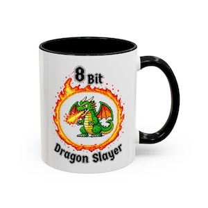 Peut inclure: Mug en céramique blanche avec intérieur et anse noirs. Il présente un dragon vert pixélisé crachant du feu, entouré d'un anneau orange flamboyant. Le texte "8 Bit Dragon Slayer" est affiché sur le mug.