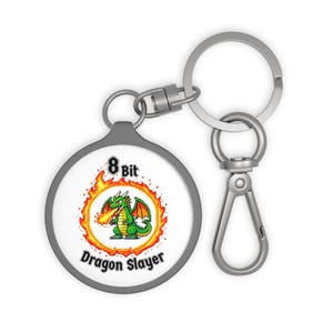 May include: A round keychain with a gray border and a silver clasp. The center features a pixelated green dragon breathing fire, surrounded by a fiery orange circle. The text "8 Bit Dragon Slayer" is displayed.