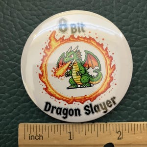 May include: A white button with the text "8 Bit Dragon Slayer". The button features a pixelated green dragon breathing fire, surrounded by a fiery orange ring. The button measures approximately 2 inches in diameter.