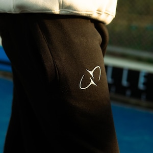 Puede incluir: Pantalones de chándal negros con un logotipo bordado en blanco en la parte superior del muslo. Los pantalones se combinan con una sudadera con capucha de color crema que descansa sobre la cintura, mostrando un diseño limpio y sencillo.