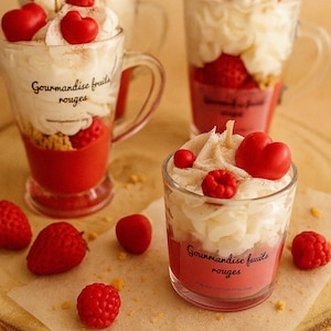 Peut inclure: Bougies en forme de desserts, présentées dans des verres transparents. Elles sont composées de couches de cire rouge et blanche, ornées de cœurs et de framboises en cire. Le texte "Gourmandise fruits rouges" est visible.