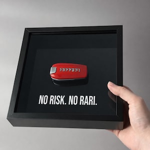May include: A red Ferrari car key displayed in a black shadow box. The key has a silver accent and the Ferrari logo. The text "NO RISK. NO RARI." is printed in white below the key.