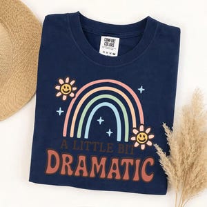 Peut inclure: Un t-shirt bleu marine à col rond avec un motif de arc-en-ciel pastel, deux fleurs souriantes et de petites étoiles. Le texte A LITTLE BIT DRAMATIC est imprimé en lettres marron. L'étiquette de la marque Comfort Colors est visible au col.