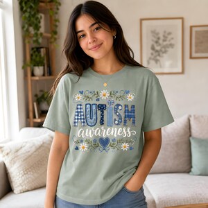 Könnte beinhalten: Ein salbeigrünes T-Shirt mit dem Schriftzug "AUTISM awareness" in einer dekorativen Schriftart. Die Buchstaben sind mit blau-weißem Karomuster und Blumenmustern gefüllt, umgeben von weißen Gänseblümchen-Illustrationen und kleinen blauen Herzen.