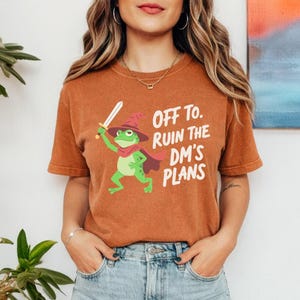 Peut inclure: T-shirt orange avec un dessin d'une grenouille magicien tenant une épée. La grenouille porte un chapeau et une cape rouges. Le texte sur le t-shirt dit "OFF TO RUIN THE DM'S PLANS".