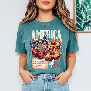 May include: Teal t-shirt with the word "AMERICA" in white block letters. The shirt features a patriotic design with cherries, pie slices, and a bow with a stars and stripes pattern. The text "LAND OF THE FREE, HOME OF THE BRAVE, Est 1776" is also included.