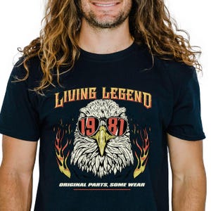 May include: Navy blue t-shirt with a graphic of an eagle wearing sunglasses and the text "LIVING LEGEND 1981". The eagle has a white head and yellow beak, with flames on either side. The text "ORIGINAL PARTS, SOME WEAR" is below.