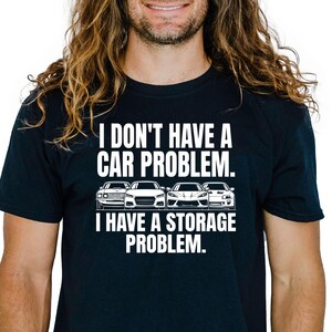 May include: Black t-shirt with the white text "I DON'T HAVE A CAR PROBLEM. I HAVE A STORAGE PROBLEM." Below the text are illustrations of four different cars. The shirt is a crew neck.