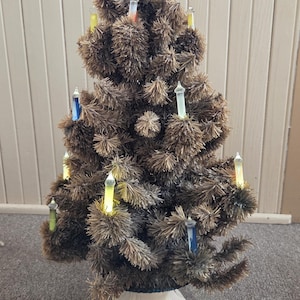 May include: A small, brown artificial Christmas tree with colorful candle-shaped lights. The tree has a white base and a clear star on top. The tree is on a gray carpet.