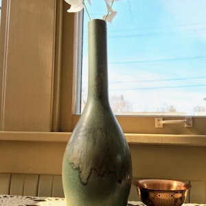 May include: A tall, green ceramic vase with a slender neck and bulbous body, holding white flowers. A small, ornate copper bowl sits beside the vase on a lace doily, with a window and blue sky in the background.
