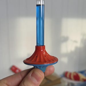 Może przedstawiać: Zabytkowy szklany instrument z niebieskim płynem w środku. Instrument ma czerwoną i niebieską podstawę. Przezroczysta szklana rurka zwęża się ku górze. Instrument jest trzymany w dłoni osoby.