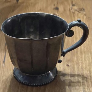 Könnte beinhalten: Eine silberfarbene Tasse mit Henkel und dekorativem Fuß. Die Tasse hat ein geriffeltes Design am Körper und einen abgerundeten Henkel mit dekorativen Elementen. Der Fuß ist mit einem Perlenmuster verziert. Die Tasse ist wahrscheinlich aus Metall.