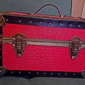 Puede incluir: Un baúl rojo de estilo vintage con ribetes negros y herrajes color latón. El baúl tiene un patrón texturizado, similar al de un cocodrilo, en la parte delantera y los laterales. Hay un asa marrón en la parte superior.