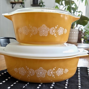 Puede incluir: Dos fuentes de horno Pyrex vintage de color amarillo mostaza con diseños florales blancos. Una fuente está apilada sobre la otra, con una tapa blanca entre ellas. Las fuentes están sobre una mesa de madera con un paño a rayas blancas y negras.