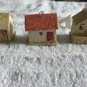 Puede incluir: Tres casas de papel en miniatura con diferentes colores y diseños de tejado. Las casas son de color beige con detalles rojos y negros, y cada una tiene un pequeño lazo de metal para colgar. Probablemente son adornos o artículos decorativos.