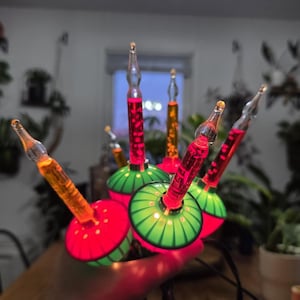 Puede incluir: Un grupo de luces navideñas vintage con bases rojas y verdes y bombillas transparentes y puntiagudas. Las bombillas contienen líquido rojo y naranja. Las luces están conectadas por un cable negro, para decoraciones festivas.