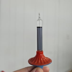 Może przedstawiać: Wintage szklana rurka z ciemnym płynem, osadzona na czerwono-niebieskiej plastikowej podstawie. Rurka jest przezroczysta z wąską końcówką. Podstawa jest z czerwonego i niebieskiego plastiku.