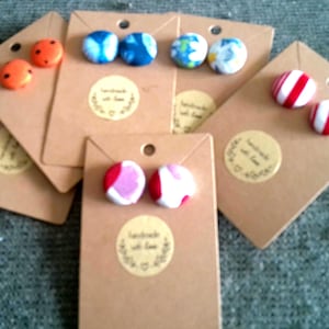 Puede incluir: Una colección de pendientes de botón hechos a mano con varios diseños, incluyendo lunares naranjas, estampados florales y rayas rojas y blancas. Cada par está montado en una tarjeta marrón con una pegatina circular que dice "handmade with love".