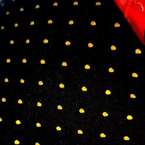 May include: A black fabric surface featuring a repeating pattern of small, circular gold-colored dots arranged in a grid. A portion of a bright red fabric edge is visible in the upper right corner, contrasting with the dark, dotted material.