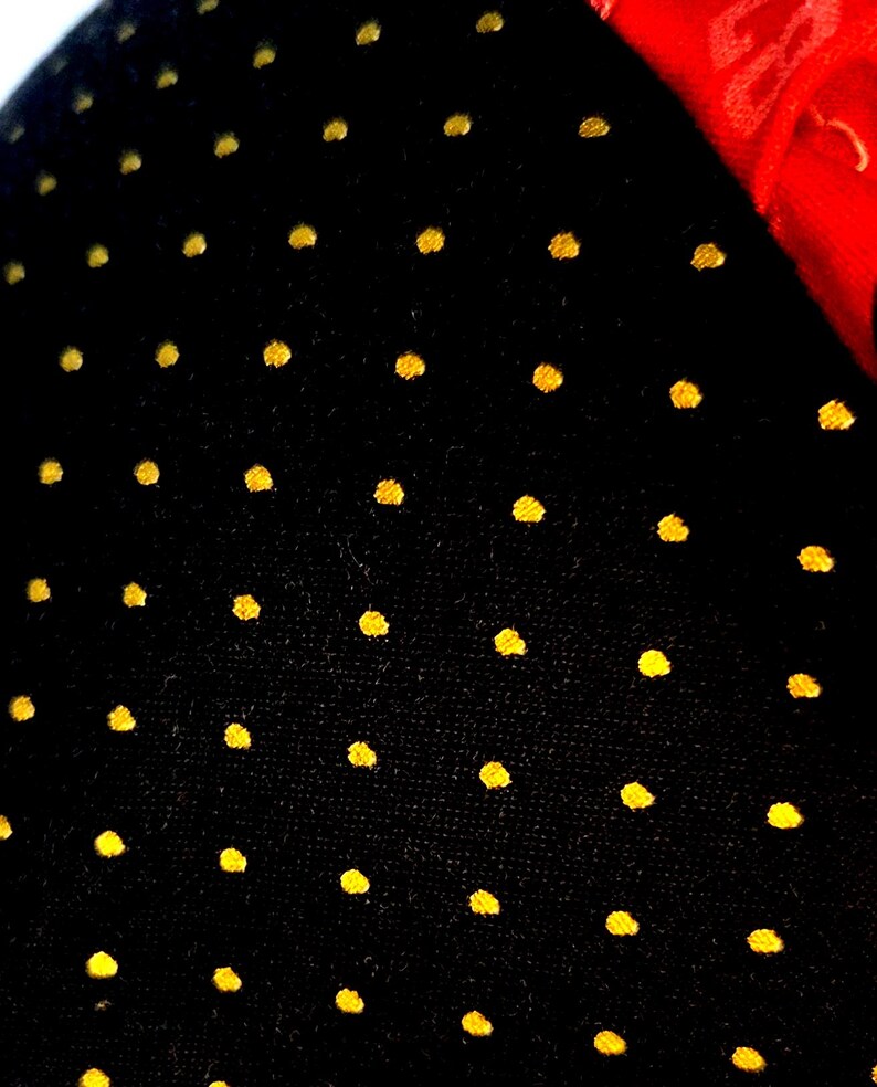 May include: A black fabric surface featuring a repeating pattern of small, circular gold-colored dots arranged in a grid. A portion of a bright red fabric edge is visible in the upper right corner, contrasting with the dark, dotted material.