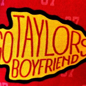 May include: A red fabric background featuring a repeating pattern of the number 87 in a lighter shade of red. Centered is a yellow arrowhead-shaped patch with a black border, embroidered with the red text GO TAYLORS BOYFRIEND.