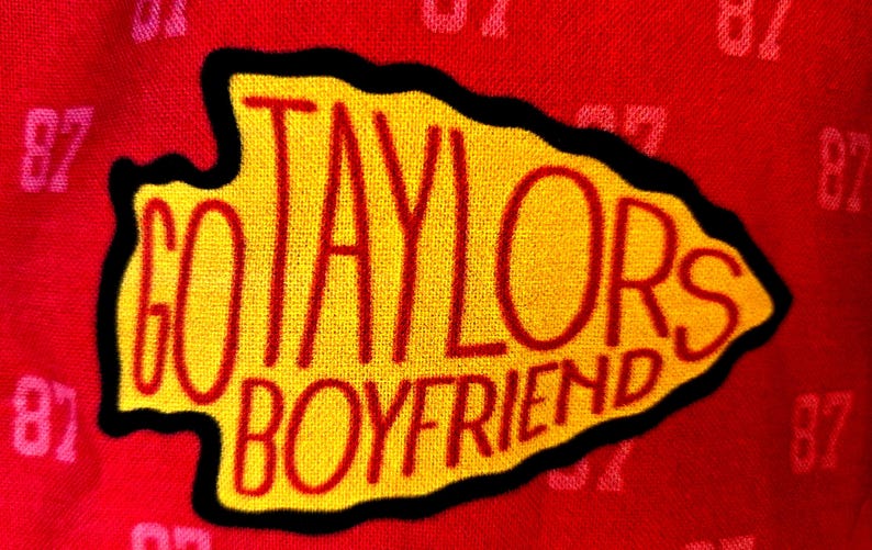 May include: A red fabric background featuring a repeating pattern of the number 87 in a lighter shade of red. Centered is a yellow arrowhead-shaped patch with a black border, embroidered with the red text GO TAYLORS BOYFRIEND.