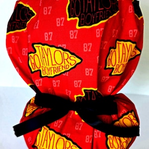 May include: A red scrub cap featuring a repeating pattern of the text "GO TAYLORS BOYFRIEND" inside yellow arrowhead shapes and the number "87" in white. The cap includes a black ribbon tie at the back to secure the fabric in a gathered style.