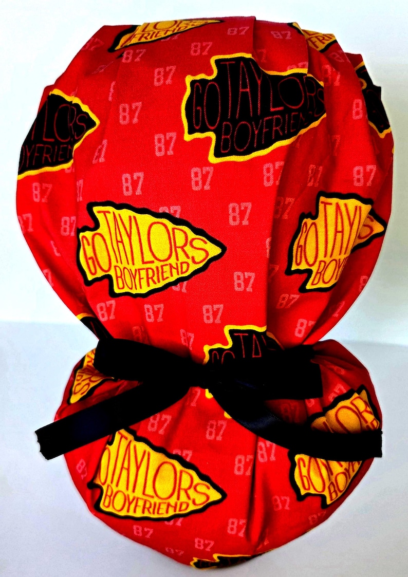 May include: A red scrub cap featuring a repeating pattern of the text "GO TAYLORS BOYFRIEND" inside yellow arrowhead shapes and the number "87" in white. The cap includes a black ribbon tie at the back to secure the fabric in a gathered style.