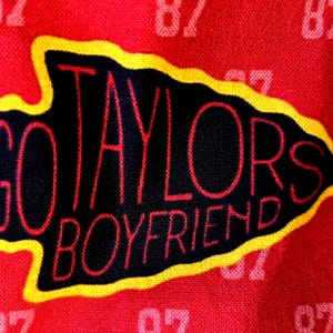 May include: A red fabric featuring a repeating pattern of the number 87 in a lighter shade of red. Centered is a black arrowhead shape with a yellow border, containing the red text GO TAYLORS BOYFRIEND written in a stylized, thin font.