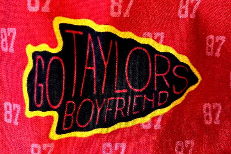 May include: A red fabric featuring a repeating pattern of the number 87 in a lighter shade of red. Centered is a black arrowhead shape with a yellow border, containing the red text GO TAYLORS BOYFRIEND written in a stylized, thin font.