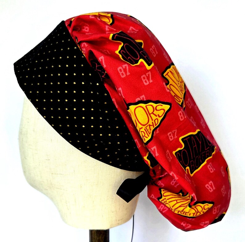 May include: A scrub cap featuring a red fabric crown with a repeating pattern of yellow and black shapes containing the text GO TAYLOR BOYFRIEND and the number 87. The front band is black with a repeating pattern of small gold dots.