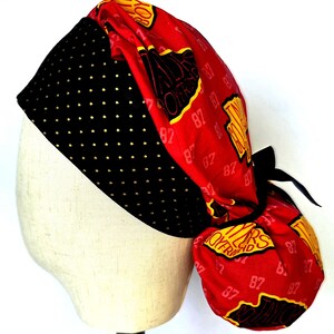 May include: A scrub cap featuring a black band with small gold dots and a red fabric section printed with the text Taylor's Boyfriend and the number 87 in yellow and black. The cap includes a black tie closure at the back to secure the fit.