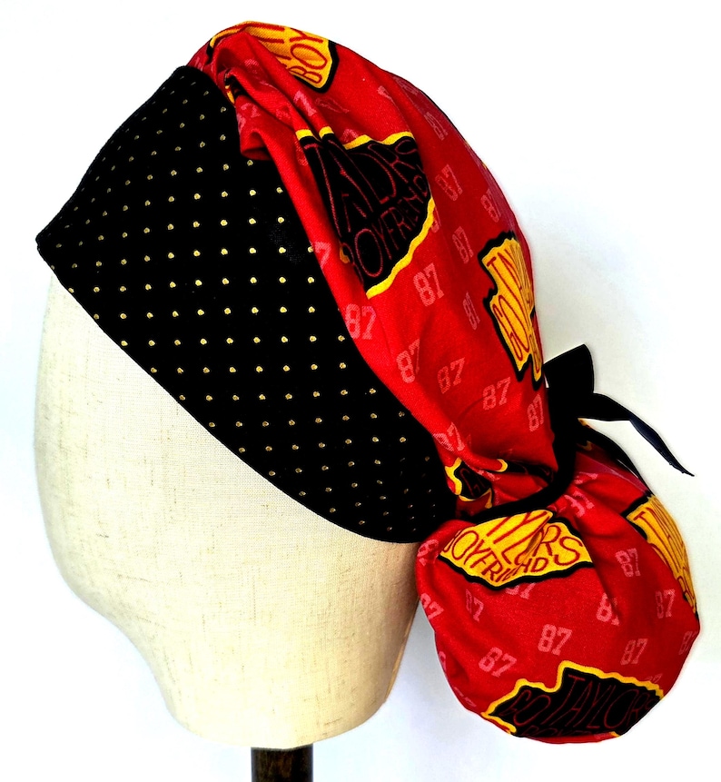 May include: A scrub cap featuring a black band with small gold dots and a red fabric section printed with the text Taylor's Boyfriend and the number 87 in yellow and black. The cap includes a black tie closure at the back to secure the fit.