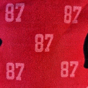 May include: A vibrant red fabric featuring a repeating pattern of the number 87 printed in a light, off-white color. The texture of the textile is visible, showing a woven surface with the numerical print distributed evenly across the material.