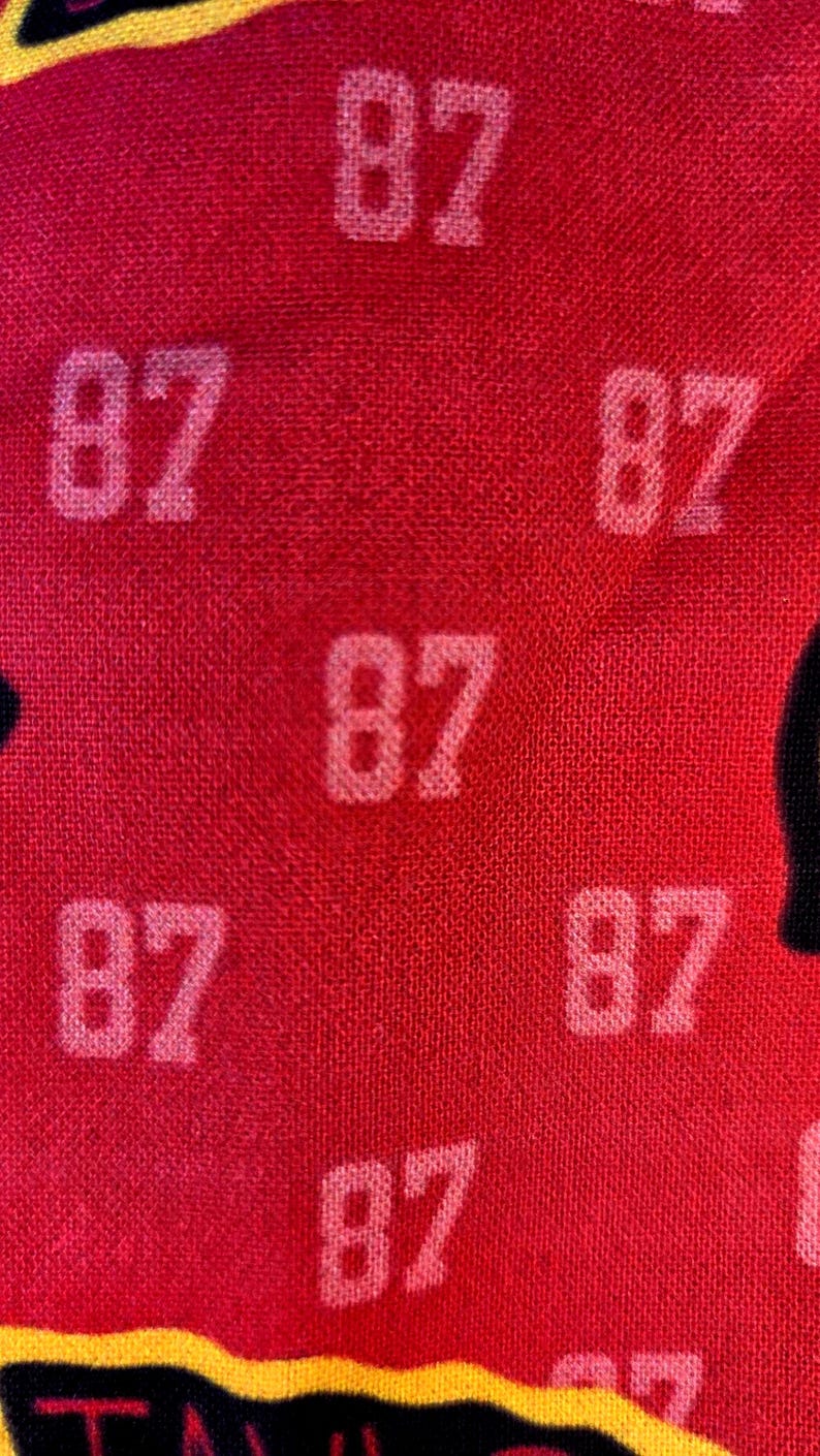 May include: A vibrant red fabric featuring a repeating pattern of the number 87 printed in a light, off-white color. The texture of the textile is visible, showing a woven surface with the numerical print distributed evenly across the material.