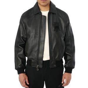 May include: Black leather bomber jacket with a collared neckline, a zipper closure, and a black emblem on the chest. The jacket has ribbed cuffs and a ribbed hem. The model is wearing a white shirt underneath and black pants.