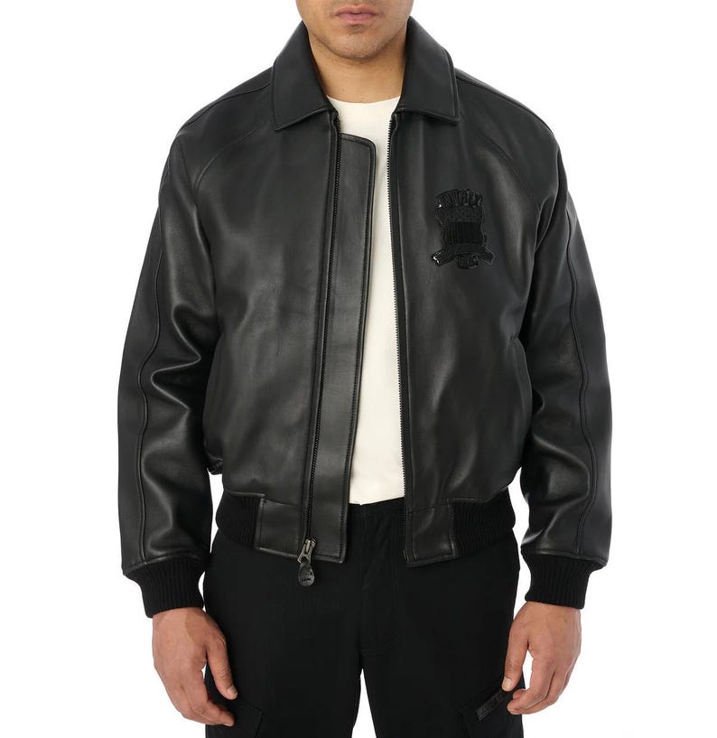 May include: Black leather bomber jacket with a collared neckline, a zipper closure, and a black emblem on the chest. The jacket has ribbed cuffs and a ribbed hem. The model is wearing a white shirt underneath and black pants.