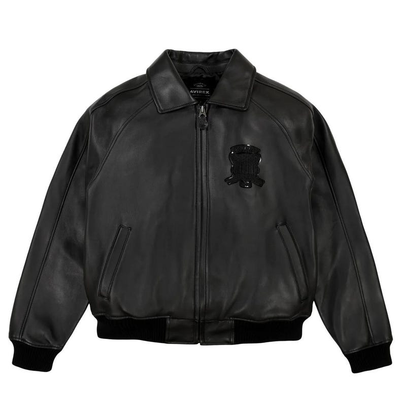 May include: Black leather bomber jacket with a zip-up front, collar, and ribbed cuffs and hem. Features a small emblem on the chest and the brand name "AVIREX" on the collar.