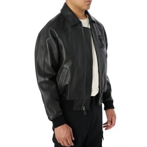 May include: Black leather bomber jacket with a collar, zipper closure, and ribbed cuffs and hem. The jacket has a classic design with a subtle logo detail on the chest. The model is wearing a white t-shirt underneath.