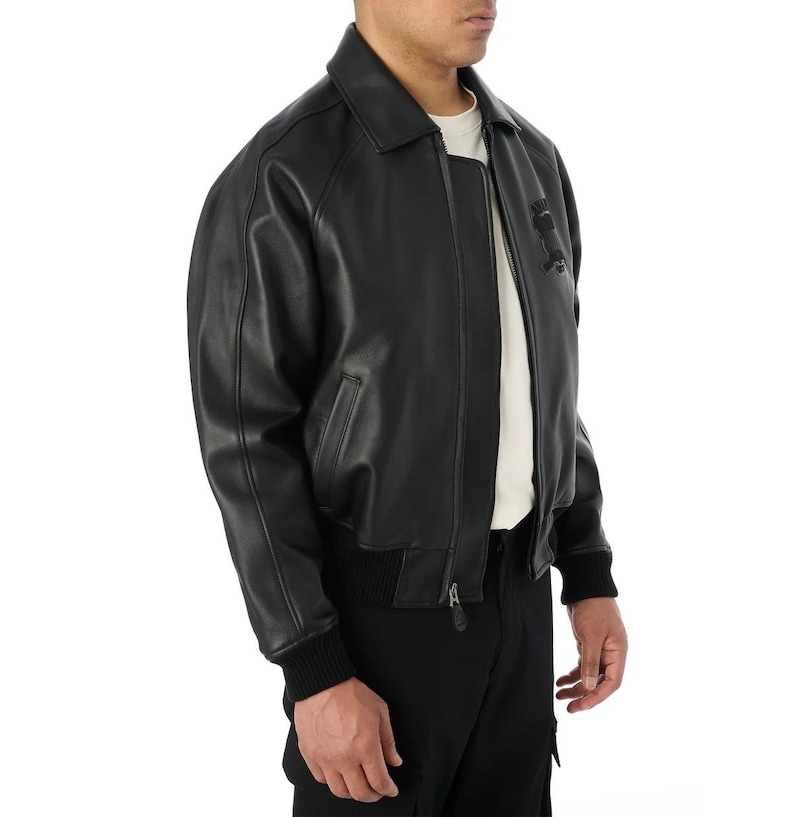 May include: Black leather bomber jacket with a collar, zipper closure, and ribbed cuffs and hem. The jacket has a classic design with a subtle logo detail on the chest. The model is wearing a white t-shirt underneath.