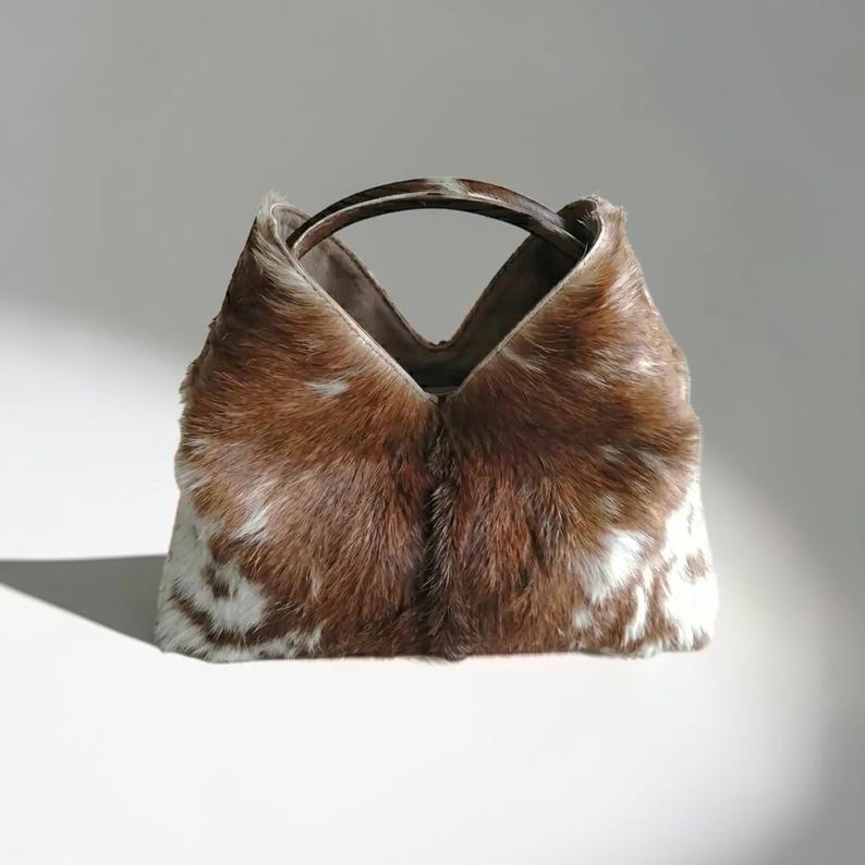 May include: A handbag crafted from brown and white cowhide with a textured, furry surface. The bag features a distinct V-shaped top opening and a single, smooth, dark brown arched handle that connects to the top edges of the bag structure.