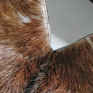 May include: A close-up view of a brown and white cowhide material featuring a natural fur texture. The hide is joined by a visible stitched seam that runs vertically, connecting two distinct sections of the patterned animal hair.