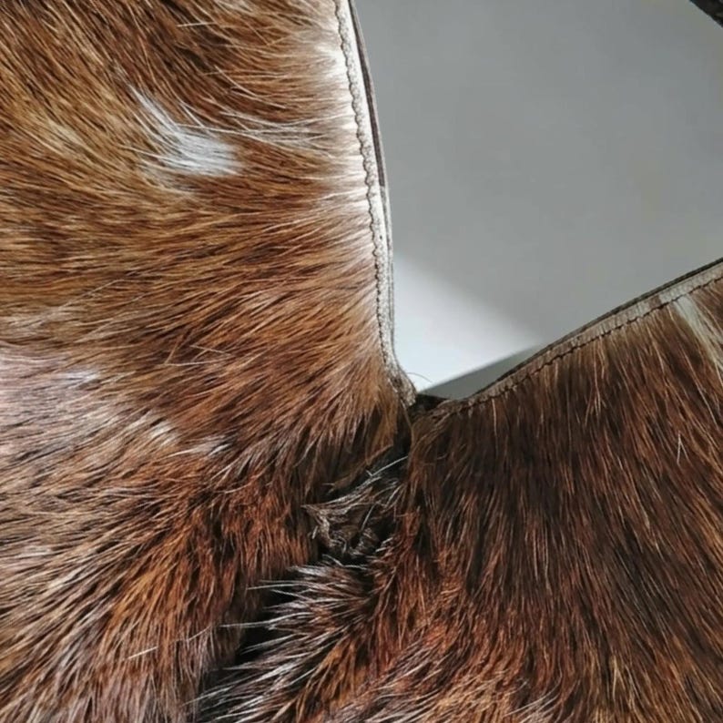 May include: A close-up view of a brown and white cowhide material featuring a natural fur texture. The hide is joined by a visible stitched seam that runs vertically, connecting two distinct sections of the patterned animal hair.