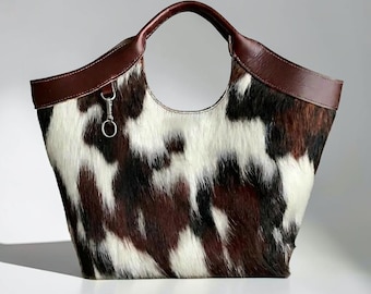 May include: A tote bag featuring a cowhide pattern with patches of white, dark brown, and black fur. The bag has a smooth dark brown leather trim along the top edge and integrated handles, with a small silver metal ring attached to the front.