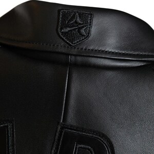 May include: Close-up of a black leather jacket collar with a small, embroidered emblem. The jacket has a classic design, with a smooth, textured surface. The emblem is a shield shape with a stylized design.